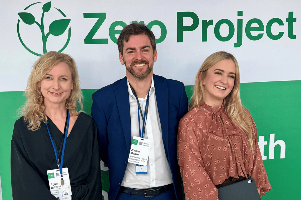 Trzy osoby stoją przed ścianką z logo Zero Project. Mają identyfikatory konferencyjne. Tło w kolorze białym i zielonym z napisem „Zero Project”.